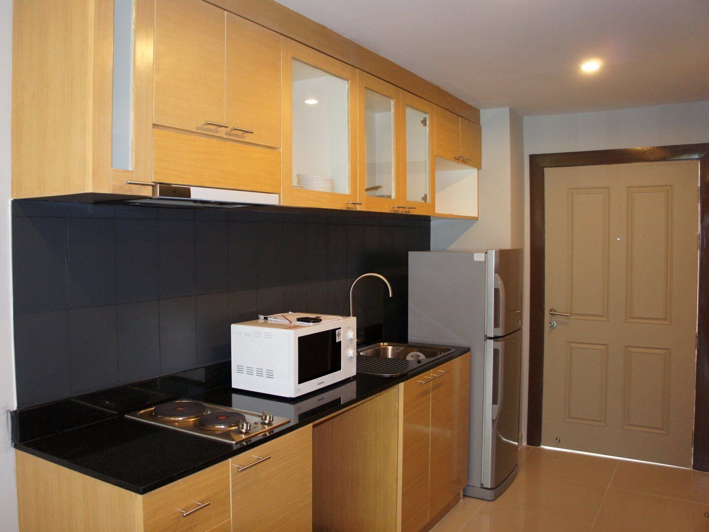 A compact kitchen with wooden cabinets, a black countertop, a white microwave, a stainless steel sink, and a refrigerator.