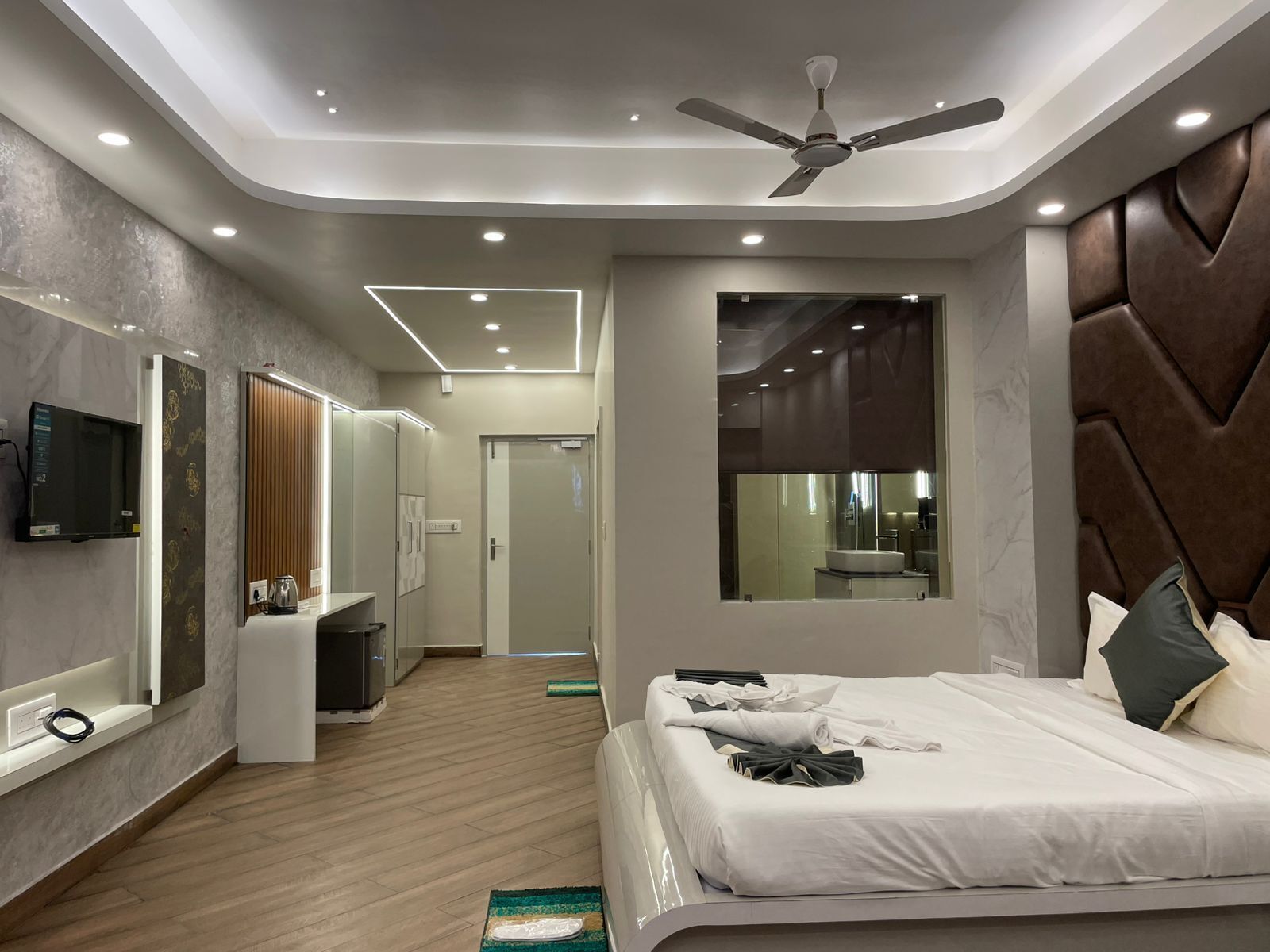 A modern hotel room with a bed, a ceiling fan, a TV, a bathroom with a mirror, and a wooden floor.