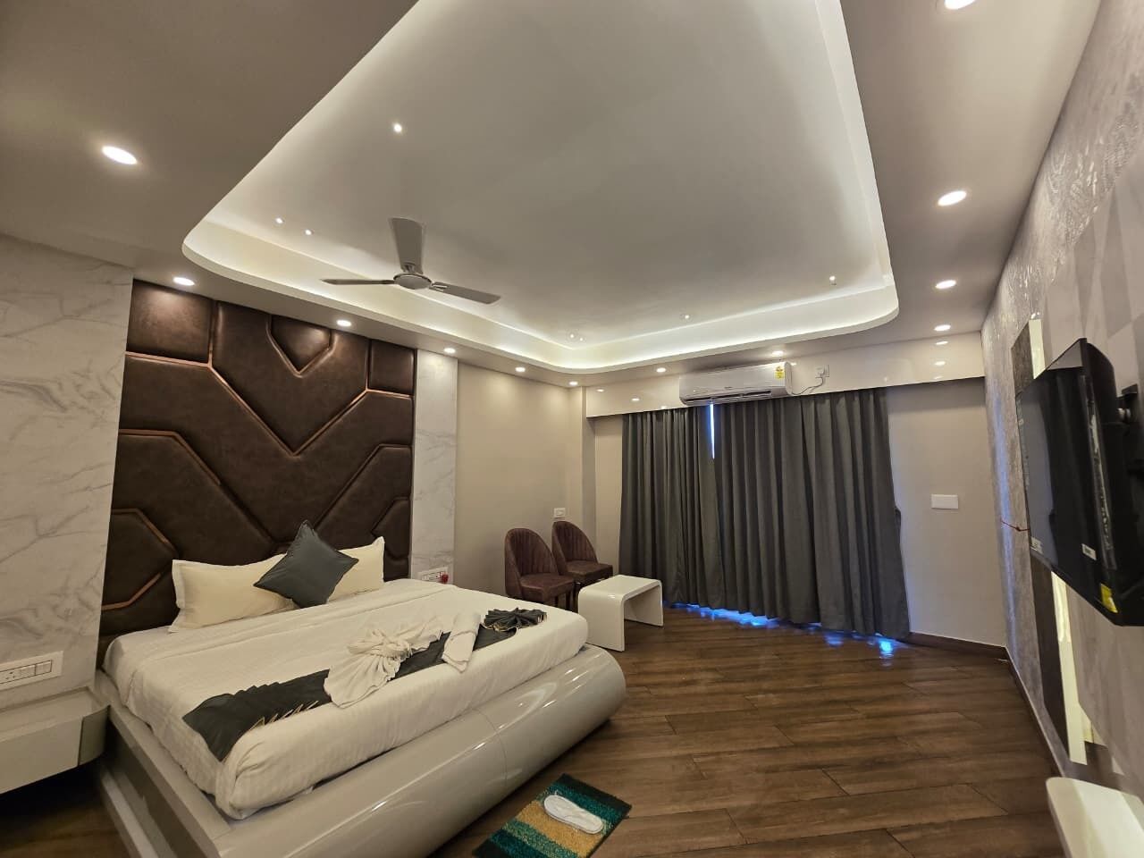 A modern hotel room with a large bed, a ceiling fan, a TV, and a seating area with chairs.