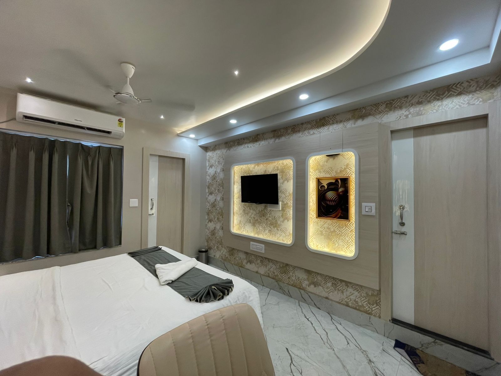 A modern hotel room with a bed, a flat-screen TV, a wall-mounted air conditioner, and a ceiling fan.