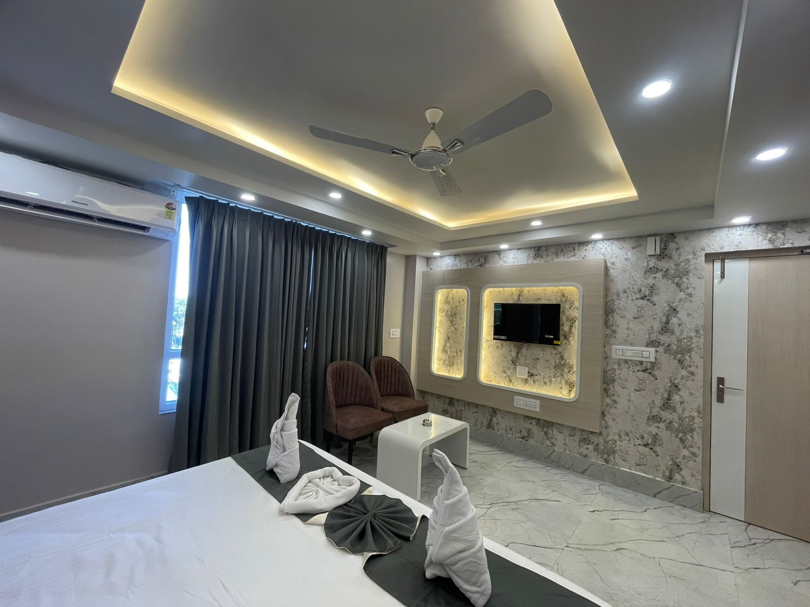 A modern hotel room with a bed, two armchairs, a small table, a ceiling fan, recessed lighting, a TV, and a door.