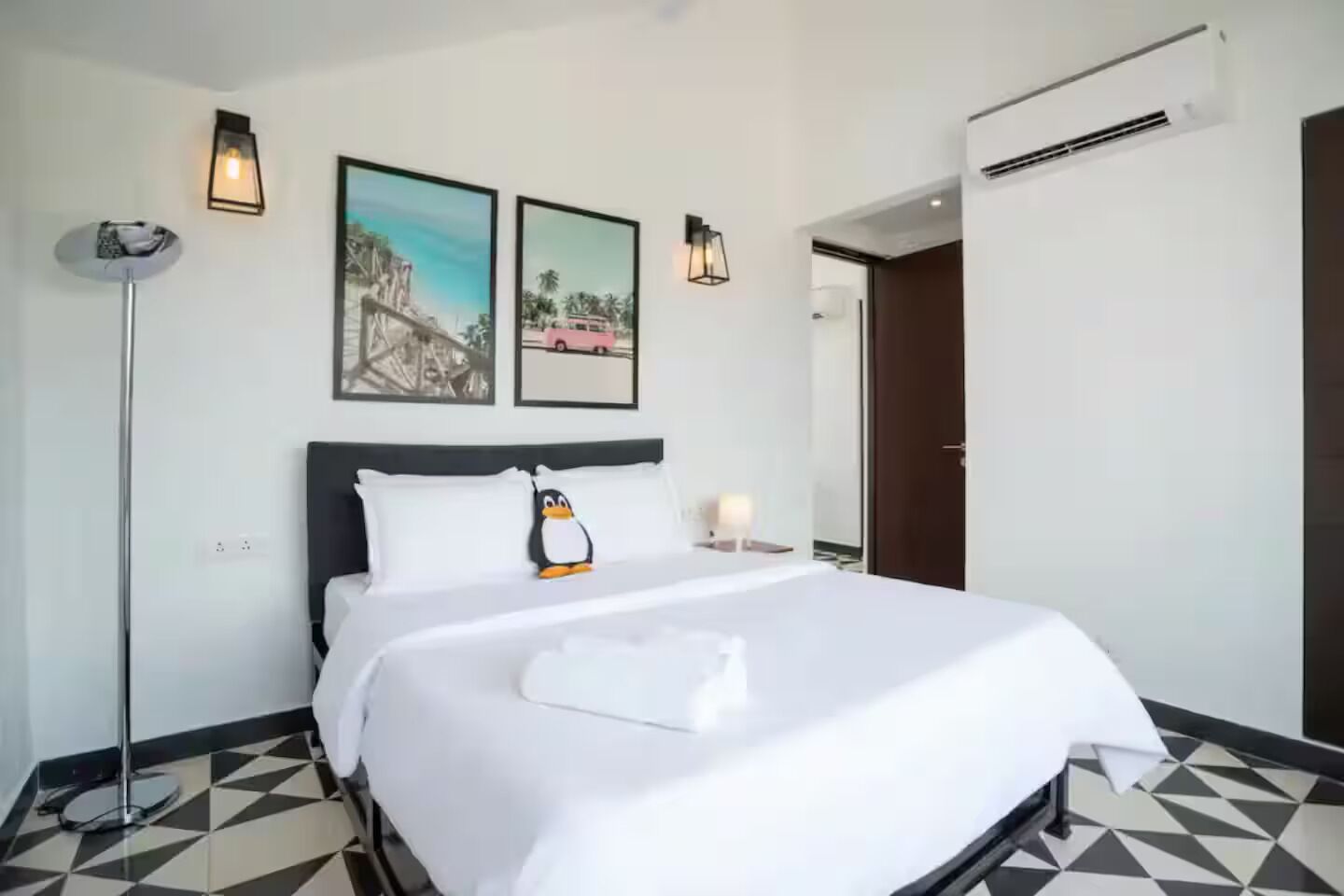 Una habitación de hotel con una cama, una lámpara de pie, dos cuadros en la pared y una puerta.