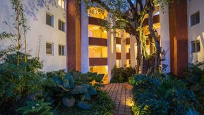 A hotel entrance with a brick pathway leading to a multi-story building, surrounded by lush greenery and trees. - The Coastal Nest - 2BHK by Da Alohas (Nerul)