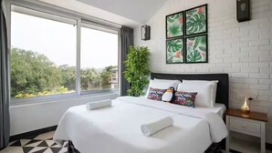 A hotel room with a large bed, a nightstand, a wall-mounted lamp, and a window with a view of trees. - The Coastal Nest - 2BHK by Da Alohas (Nerul)