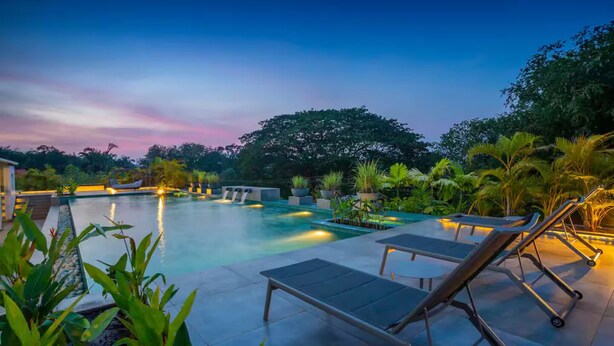 A swimming pool with lounge chairs and a waterfall feature, surrounded by lush greenery. - The Coastal Nest - 2BHK by Da Alohas (Nerul)