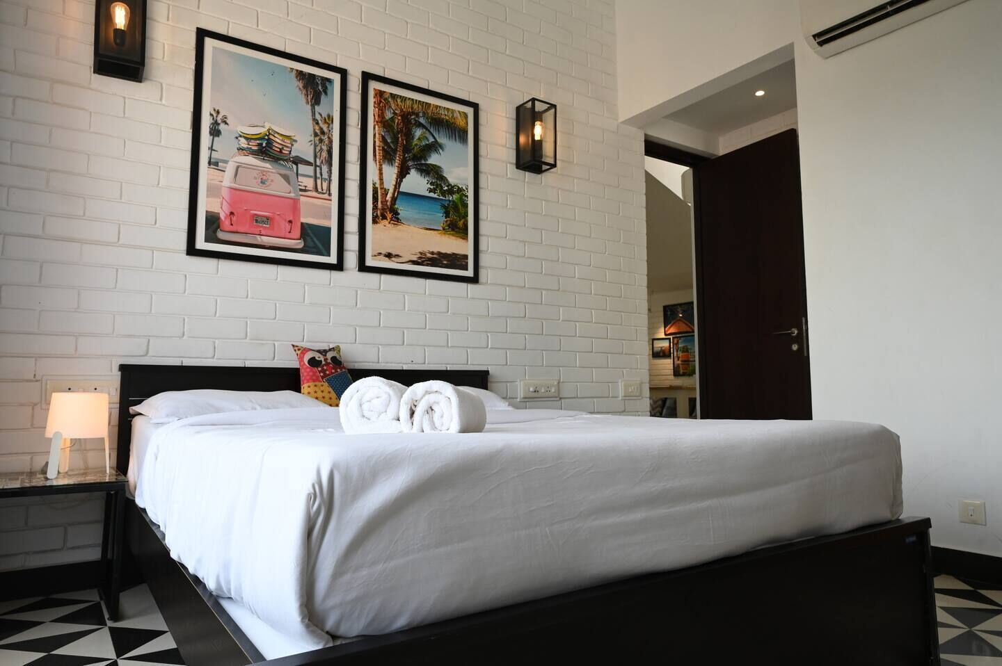 Een hotelkamer met een witte baksteenmuur, een bed met witte lakens, twee ingelijste foto's, een nachtkastje met een lamp en een zwart-wit vloertegelpatroon.