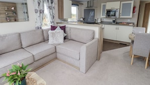 A modern living room with a beige sofa, a small table with a plant, and a kitchen area with cabinets and appliances.