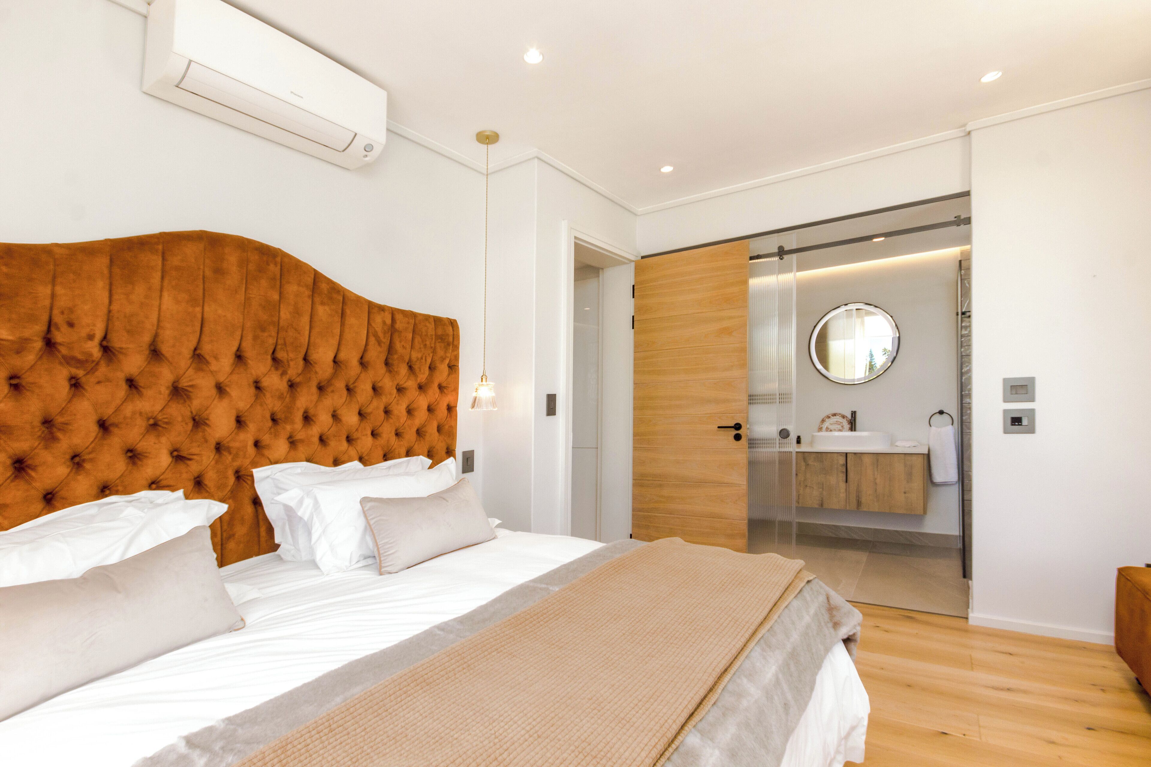 Ein modernes Schlafzimmer mit einem gepolsterten Kopfteil, einem großen Bett, einem Nachttisch und einem durch eine Glastür einsehbarer Badezimmer.