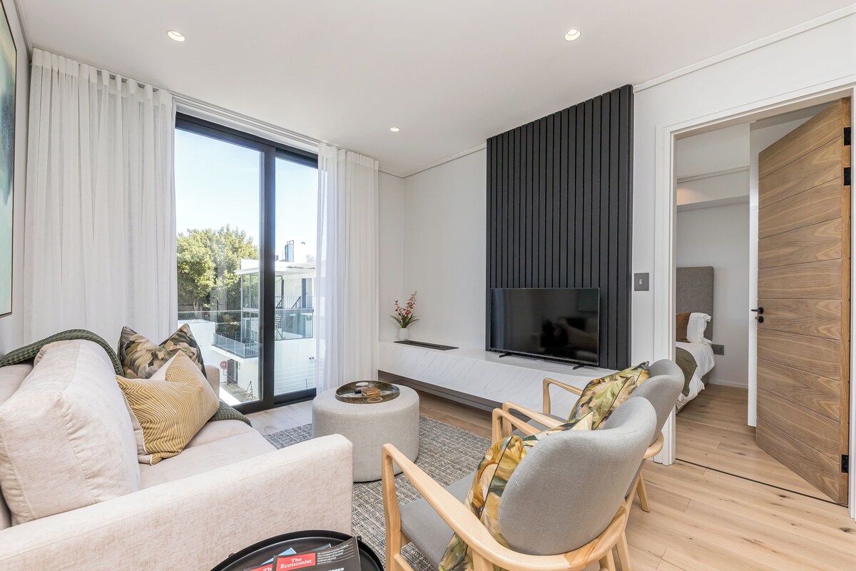 Un salon moderne comprenant un canapé, des fauteuils, une table basse, un téléviseur et une porte coulissante donnant sur une chambre.