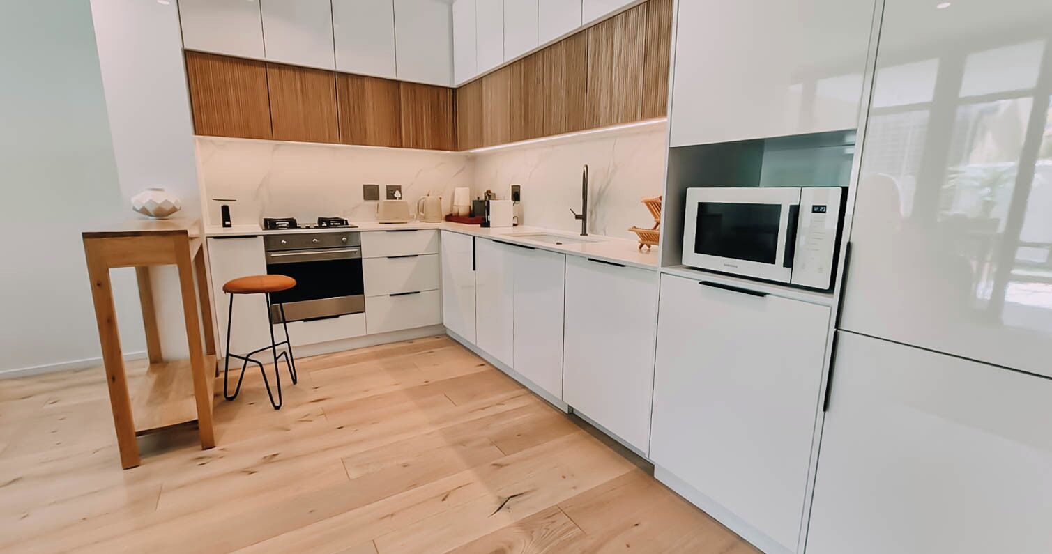 Ett modernt kök med vita skåp, en vägg med träpanel som accent, en mikrovågsugn och en träpall.