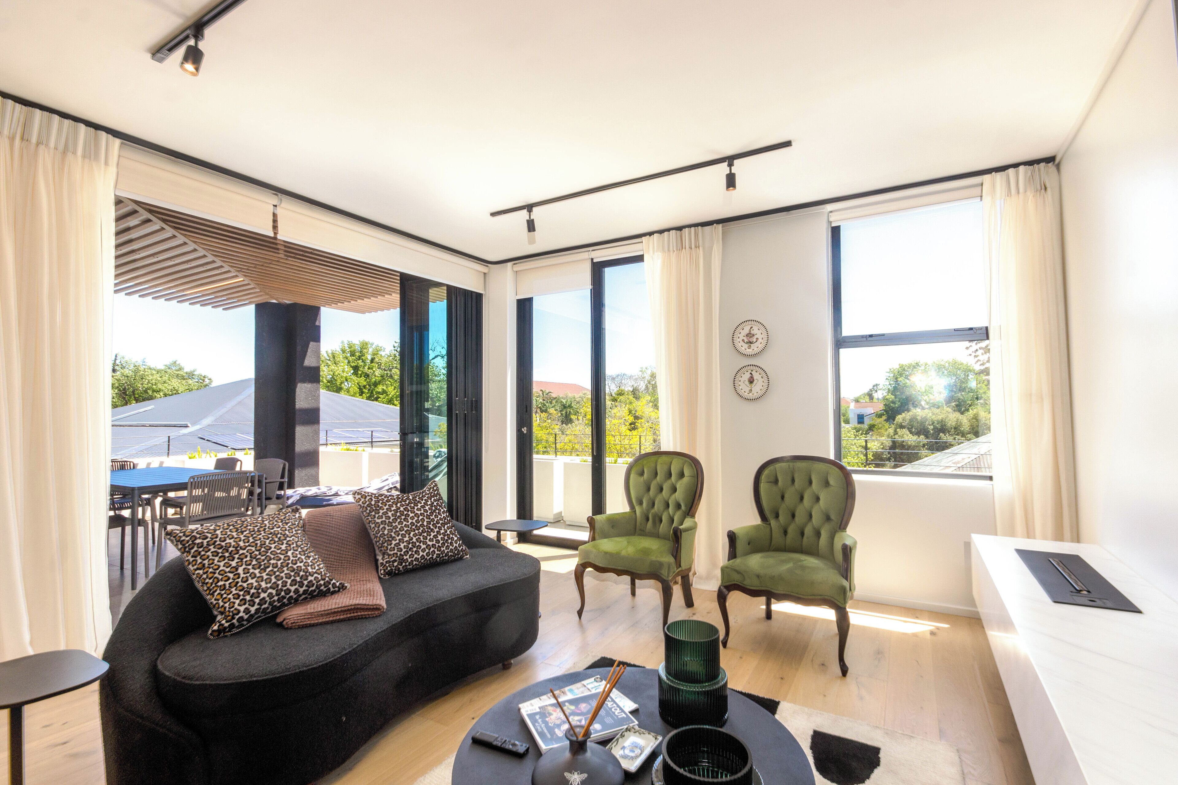 A modern living room with a sofa, two green armchairs, a dining area with a table and chairs, and large windows offering a view of the outdoors.
