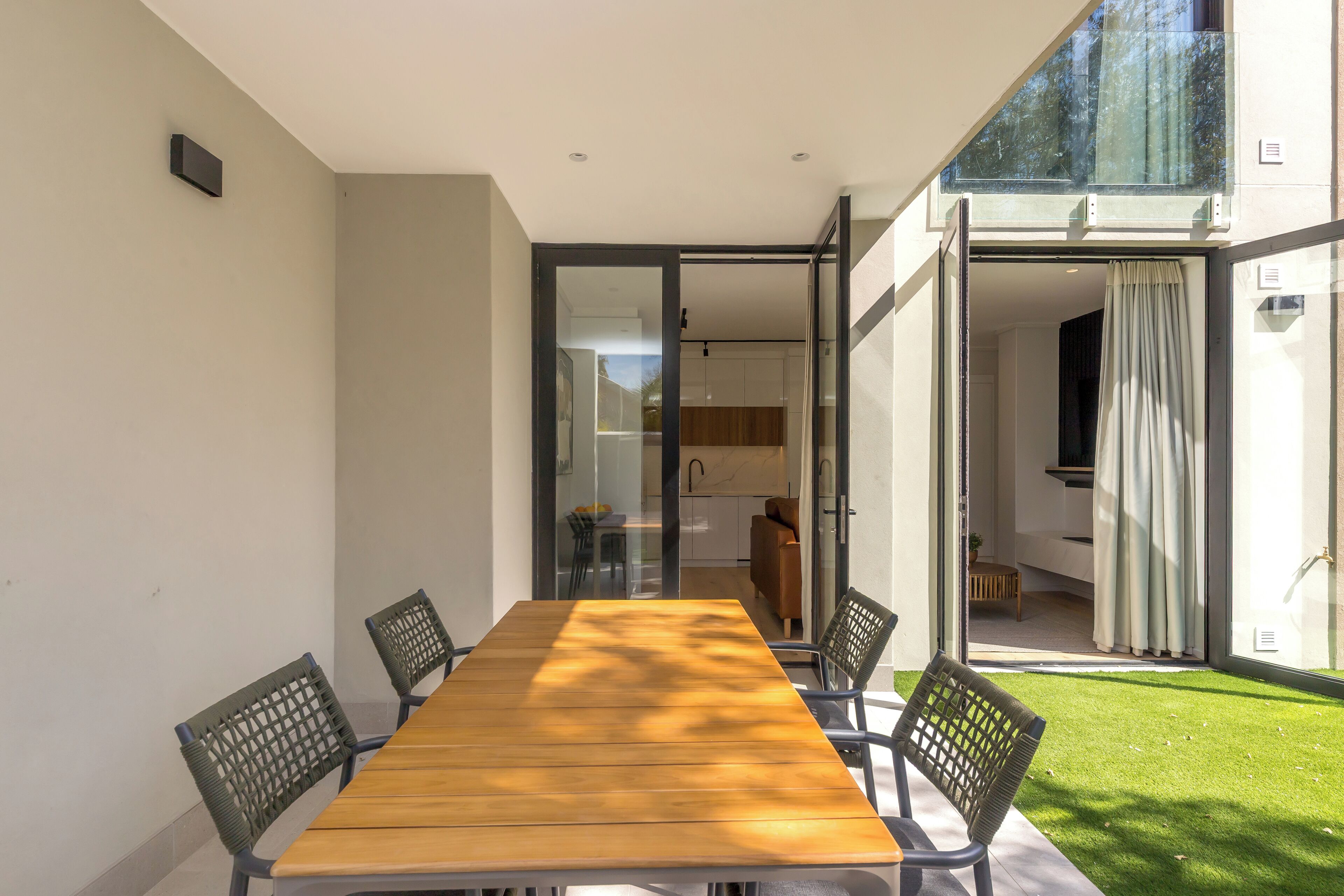 Un espace repas moderne avec une table en bois et quatre chaises, une porte vitrée donnant sur une cuisine et une vue sur une pelouse verdoyante.