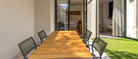 Un espace repas moderne avec une table en bois et quatre chaises, une porte vitrée donnant sur une cuisine et une vue sur une pelouse verdoyante.