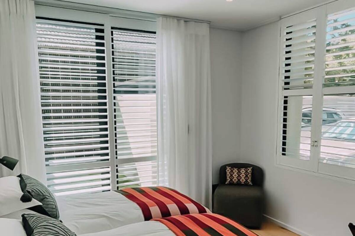 Un dormitorio con una cama grande, dos ventanas con persianas, una silla y una alfombra estampada.