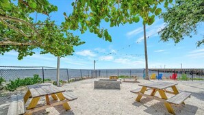 An outdoor seating area with picnic tables and benches, surrounded by a fence and trees, overlooking a body of water.
