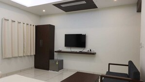 A room with a flat-screen TV mounted on the wall, a dark wooden wardrobe, a small refrigerator, a dark wooden table, and a grey sofa. - LGP Residency (Tiruchirappalli)