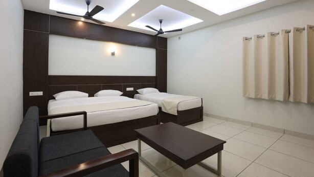 A hotel room with a large bed, two smaller beds, a sofa, a coffee table, ceiling fans, skylights, and a curtain rod. - LGP Residency (Tiruchirappalli)