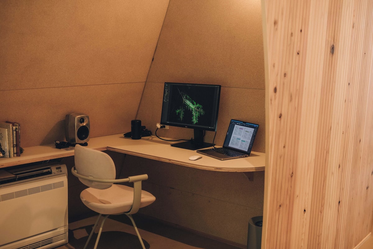 Un espacio de oficina compacto con un escritorio, dos monitores, una computadora portátil, una silla blanca, un altavoz y un calentador pequeño.