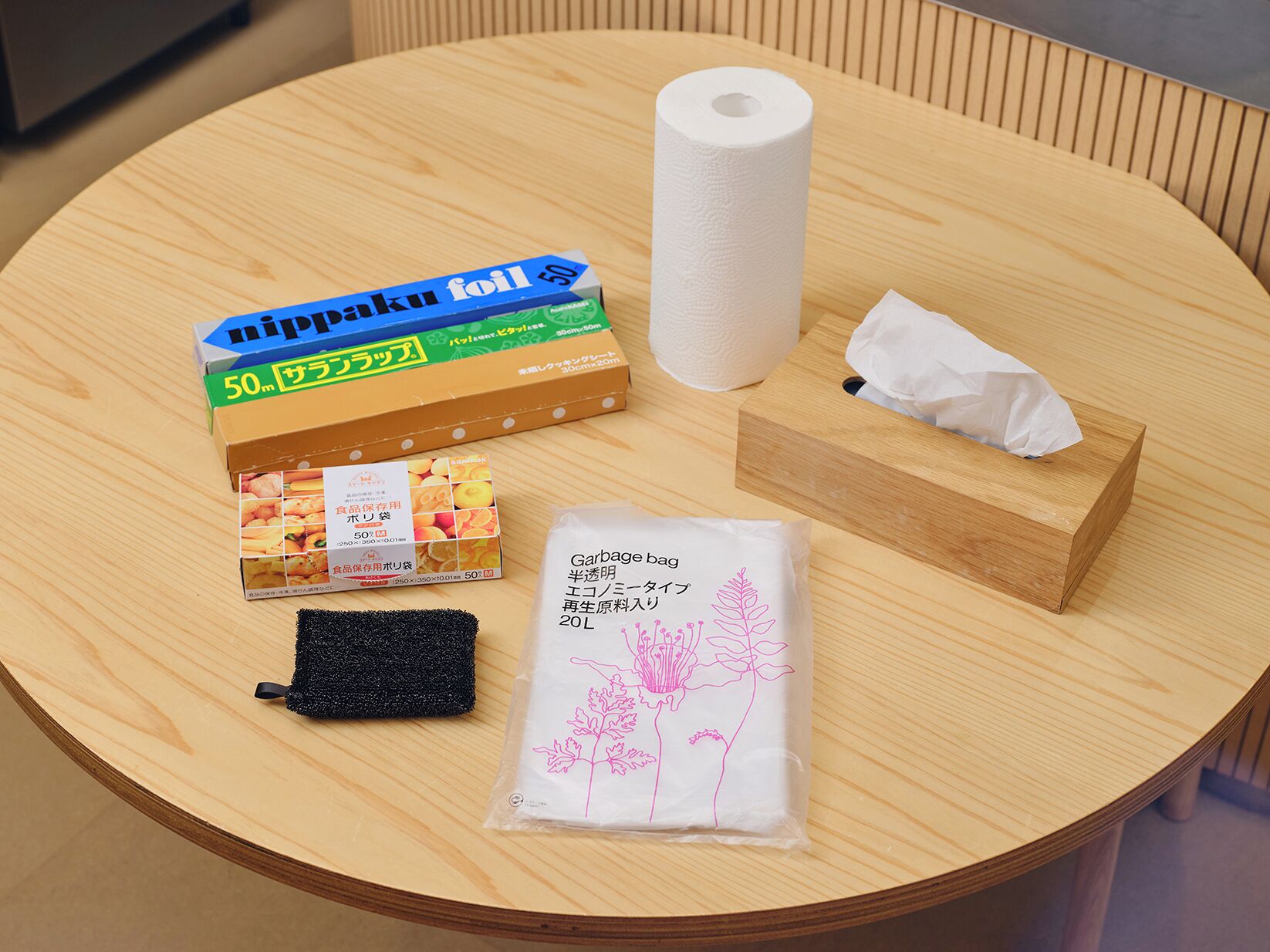 Ein runder Holztisch mit einer Toilettenpapierrolle, einer Papiertauschbox, einem schwarzen Schwamm, einem Müllbeutel und einer Packung Nippaku-Alufolie.