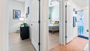 Un pasillo con puertas blancas que dan a un dormitorio con cama, ventilador de techo y baño con cortina de ducha.