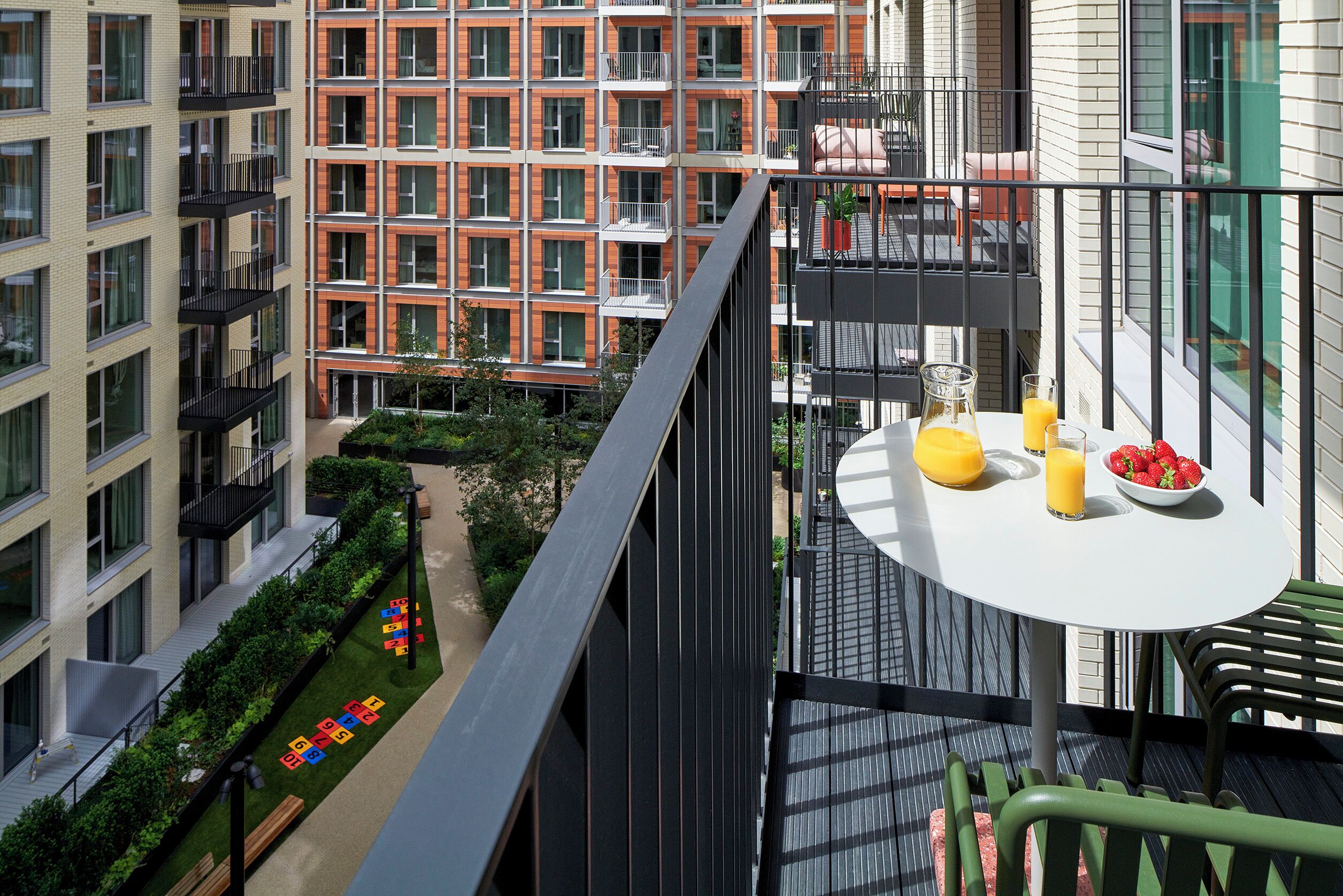 En balkon med et bord dækket til morgenmad, med udsigt over en gårdsplads med et legeområde for børn og en bygning med facade af mursten.