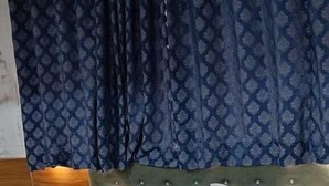 A neatly made bed with white bedding and a brown quilted cover, a wooden headboard, and a blue patterned curtain. - Shera Inn (Ahmedabad)