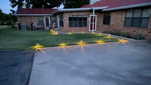 Un camino de entrada residencial con superficie de concreto, una casa de ladrillo con techo rojo y un jardín con luces decorativas.