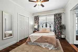Un dormitorio con cama, ventilador de techo, ventana con cortinas, espejo y puerta.