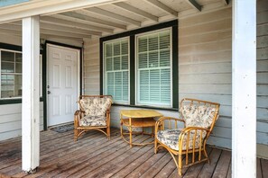A porch with two wicker chairs and a small table, a white door, and a window with shutters.