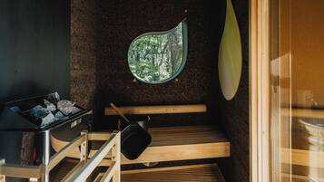 A sauna room with a wooden bench, a black bucket, and a window with a view of trees.