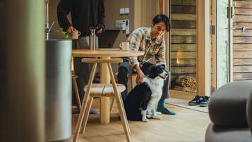 A cozy room with a wooden floor, a round table with a metal container, a cup, and a book. A person is standing by the table, and a dog is sitting on the floor.