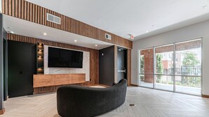 A modern living room with a curved black sofa, a large flat-screen TV mounted on a wooden wall, and a sliding glass door leading to a balcony. - Regards At Med Center 130 I Spacious 1 Bed 1 Bath (Houston)