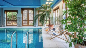 En inomhuspool med väggar av genomskinligt glas, omgiven av solstolar och krukväxter, och med stora fönster som släpper in naturligt ljus.