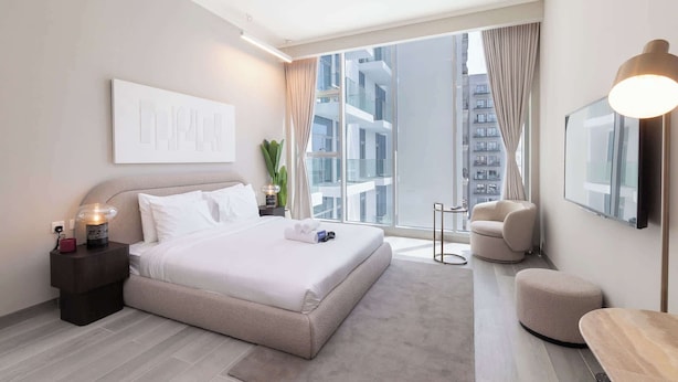 A modern hotel bedroom with a large bed, a nightstand, a chair, and a view of the city.
