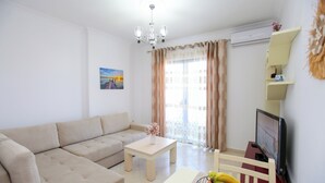 A living room with a beige sofa, a small table with a vase of flowers, a television, and a fruit bowl. - Seascape Apartments Vlore (Vlore)