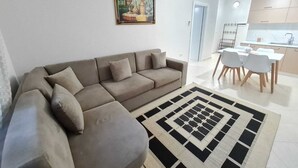 A modern living room with a beige sofa, a patterned rug, and a dining area with white chairs and a table. - Seascape Apartments Vlore (Vlore)