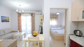 A modern, well-lit living room with a sofa, dining table, and a TV. A kitchen area is visible with a bed and a kettle. - Seascape Apartments Vlore (Vlore)