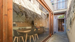 A narrow alley with a wooden table and chairs, a window with a view of a building, and a door leading to a balcony.