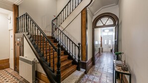 A grand staircase with wooden steps and black balusters, leading to a hallway with patterned tile flooring and a radiator on the left.