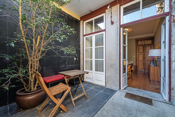 A small outdoor seating area with a wooden table and bench, a potted tree, and a doorway leading to an interior space.
