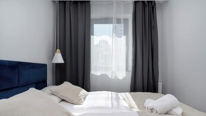 A neatly made bed with white linens and a blue headboard, a white lamp, and a view of a building through sheer curtains.