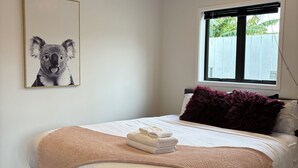 Een net opgemaakt bed met een wit en beige deken, een kunstwerk van een koala aan de muur, en een raam met uitzicht op groen.