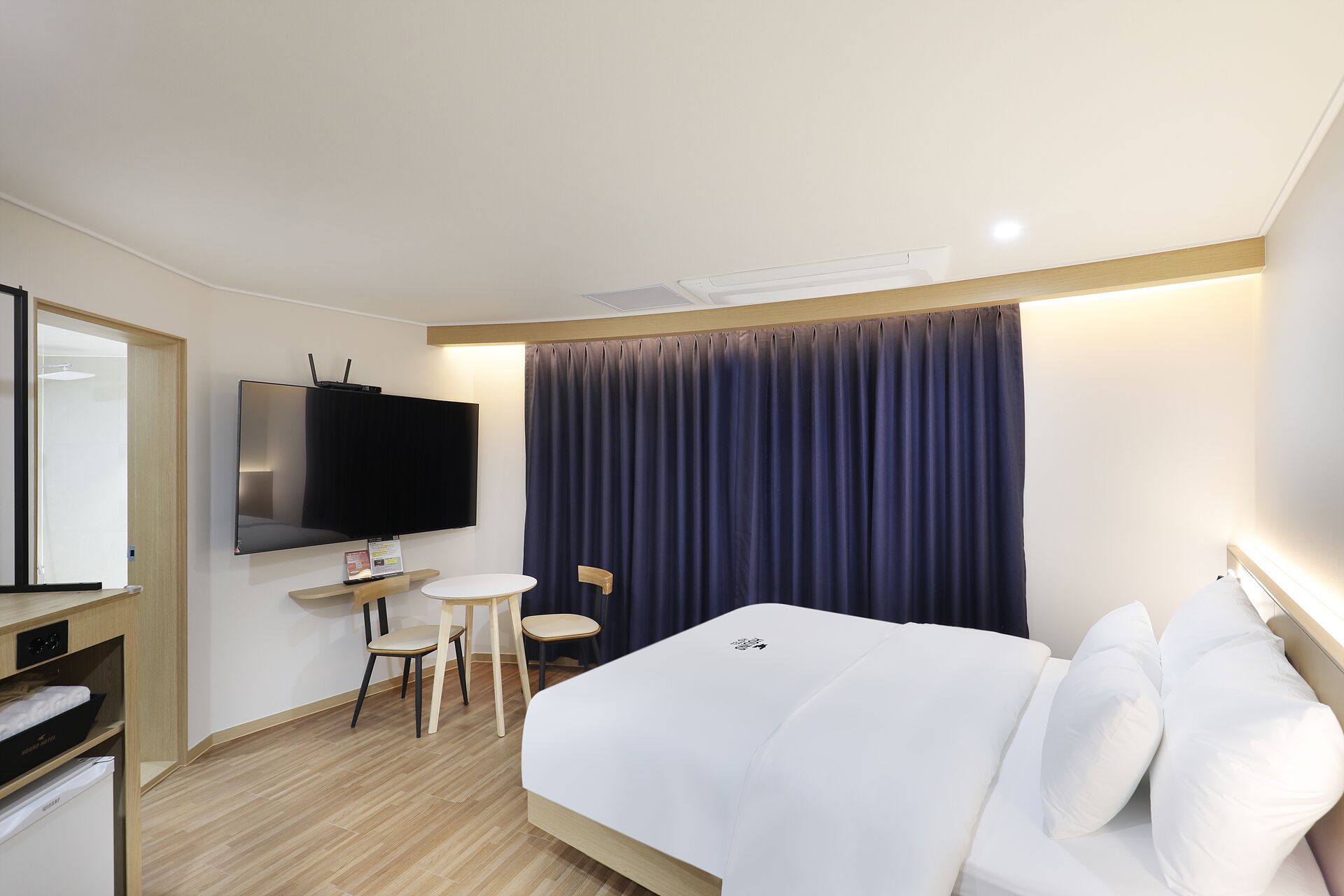 A hotel room with a bed, a desk with a chair, a flat-screen TV, and a large window with curtains.