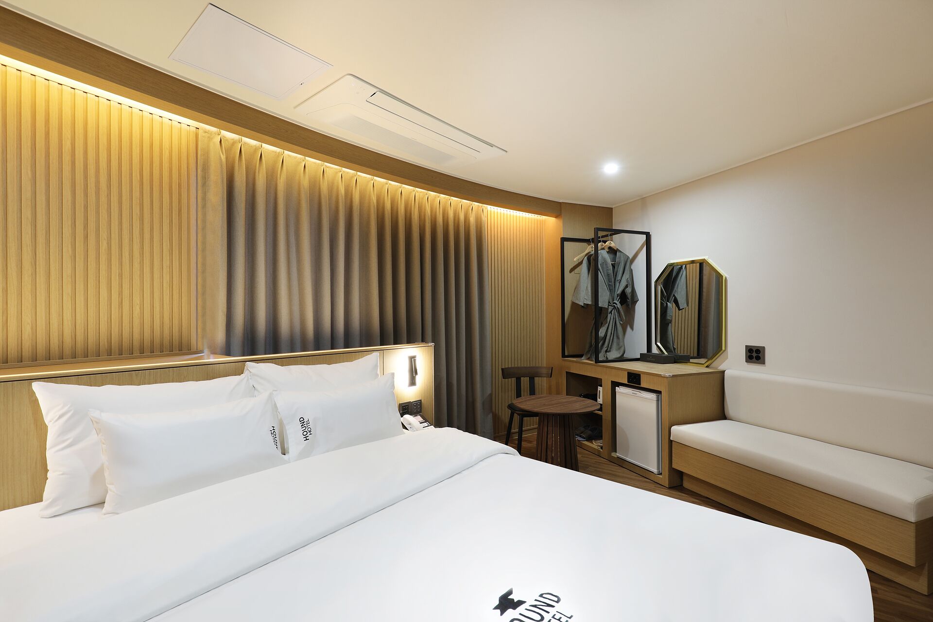 A hotel room with a bed, a seating area, a mirror, and a wall-mounted air conditioning unit.