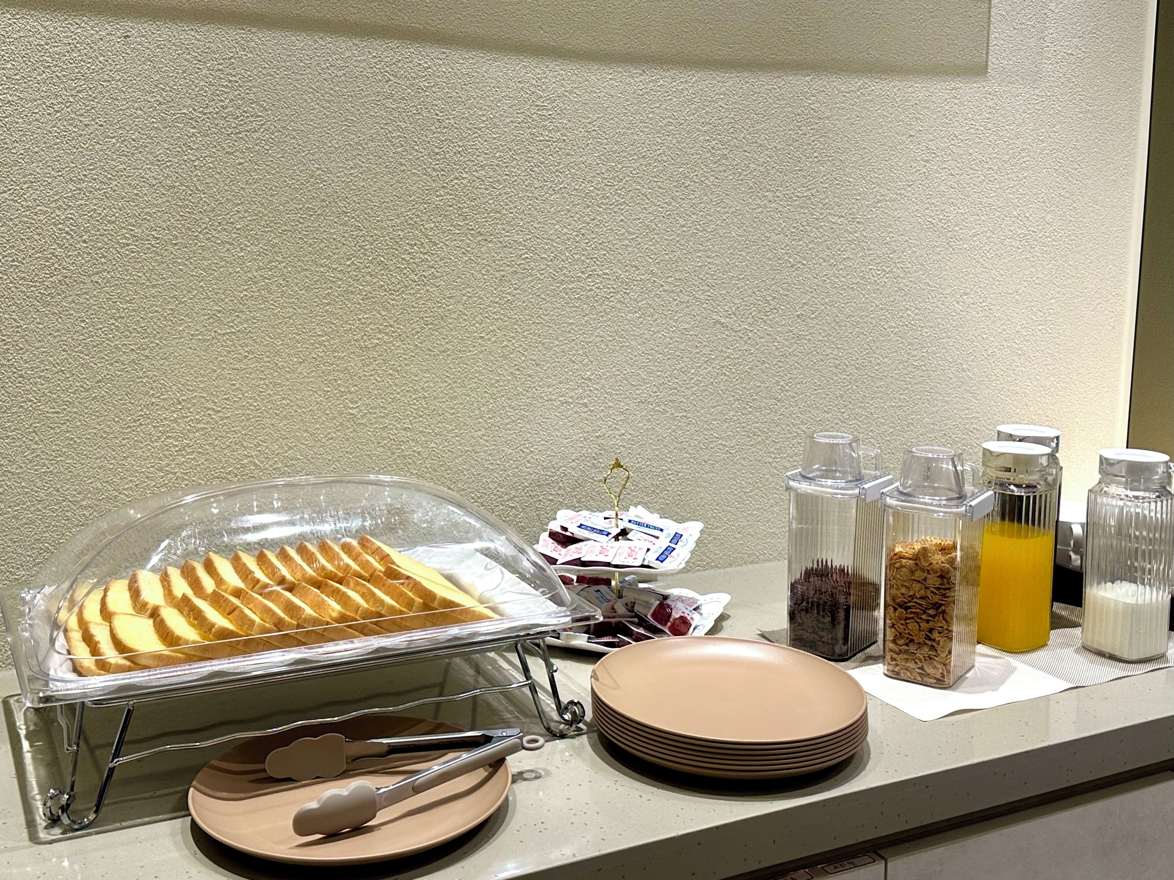 A breakfast buffet setup with a covered food display, serving tongs, plates, and various food and drink containers.