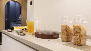 A breakfast bar with juice dispensers, a fruit bowl, and cereal containers.