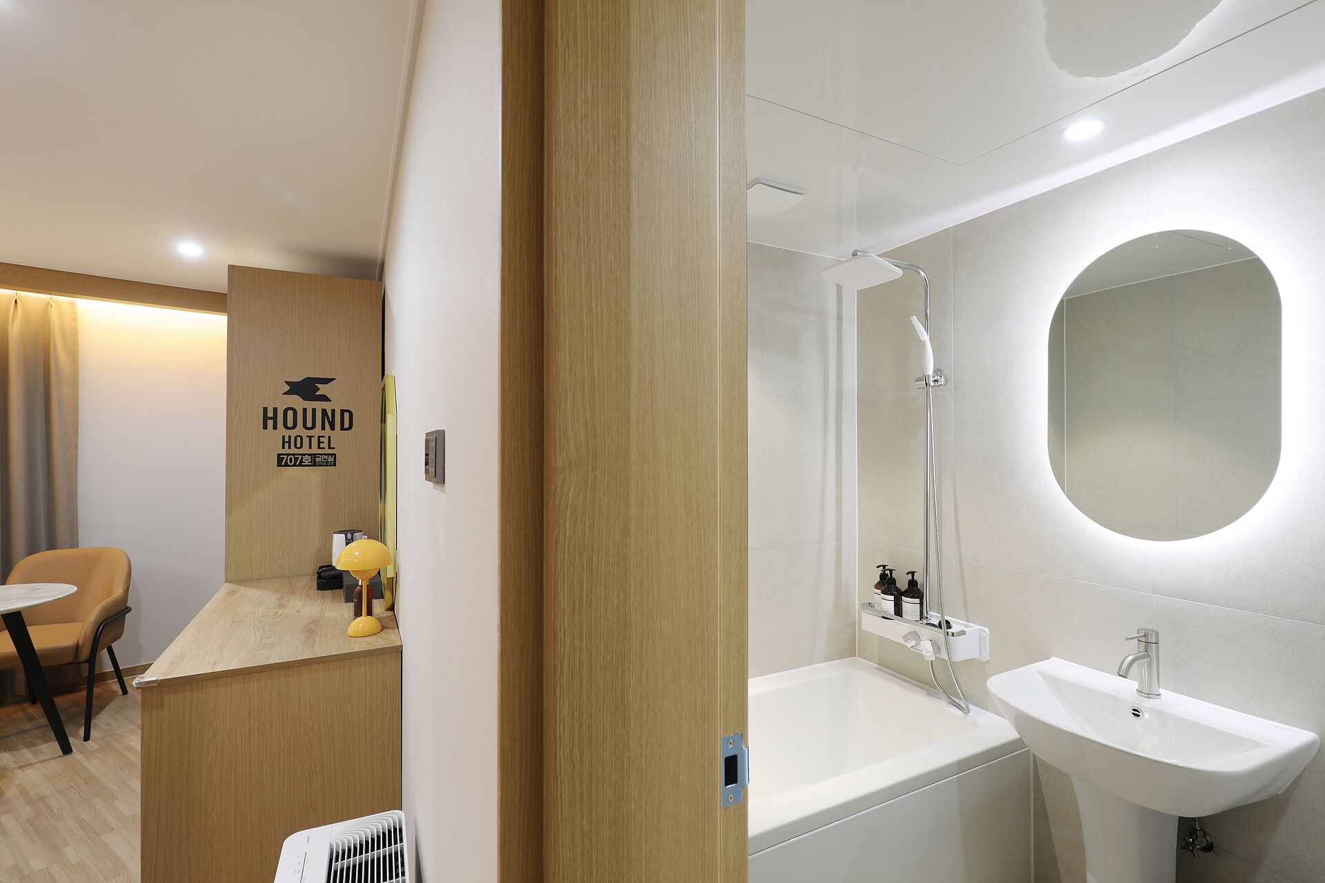 A modern hotel bathroom with a bathtub, a sink, and a shower. There is a wooden reception desk with a sign that reads "HOUND HOTEL".