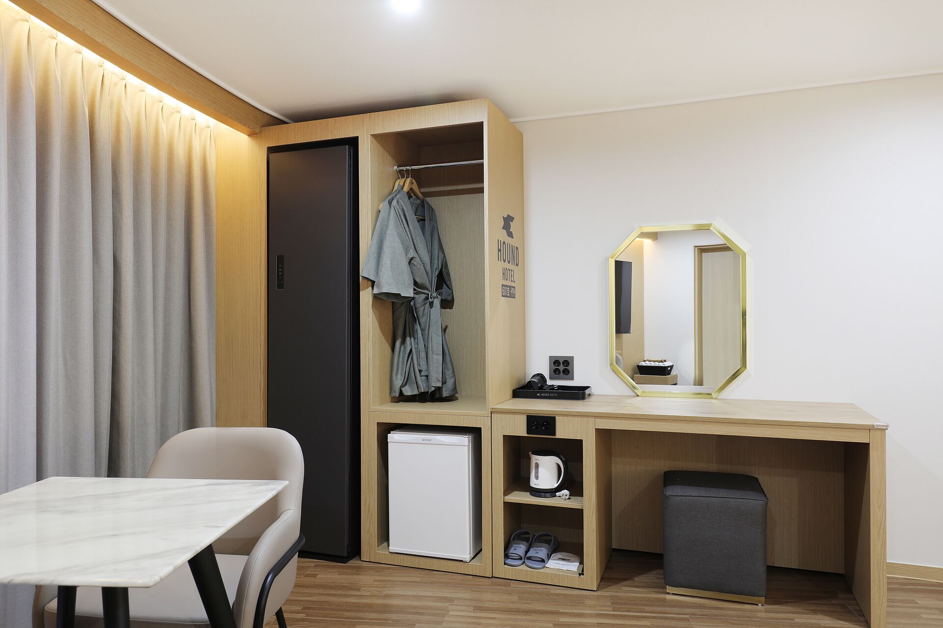 A hotel room with a desk, a chair, a wardrobe, a mirror, a small refrigerator, and a trash bin.