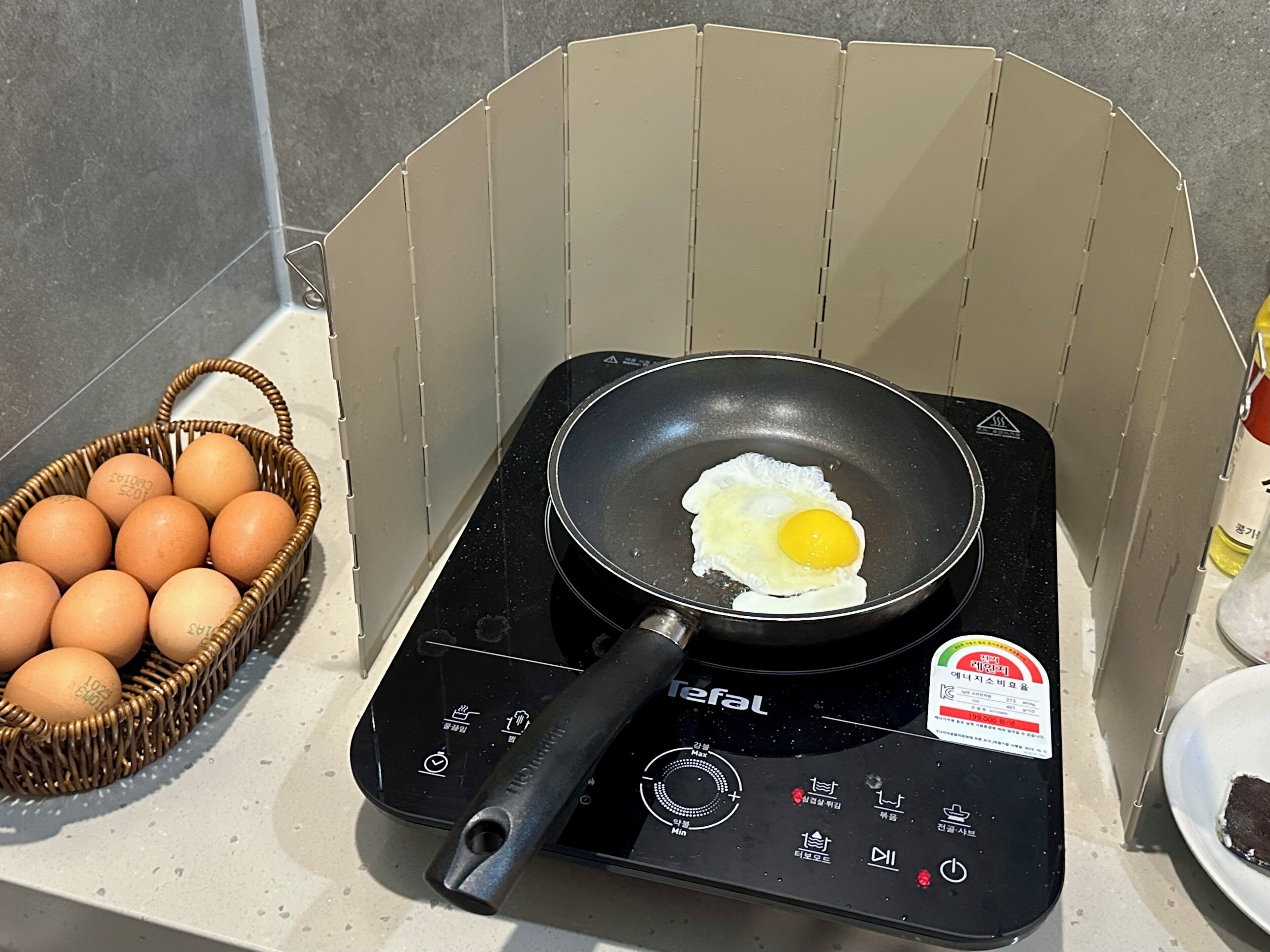 A kitchen with a frying pan on an induction cooktop, a basket of eggs, and a bottle of cooking oil.