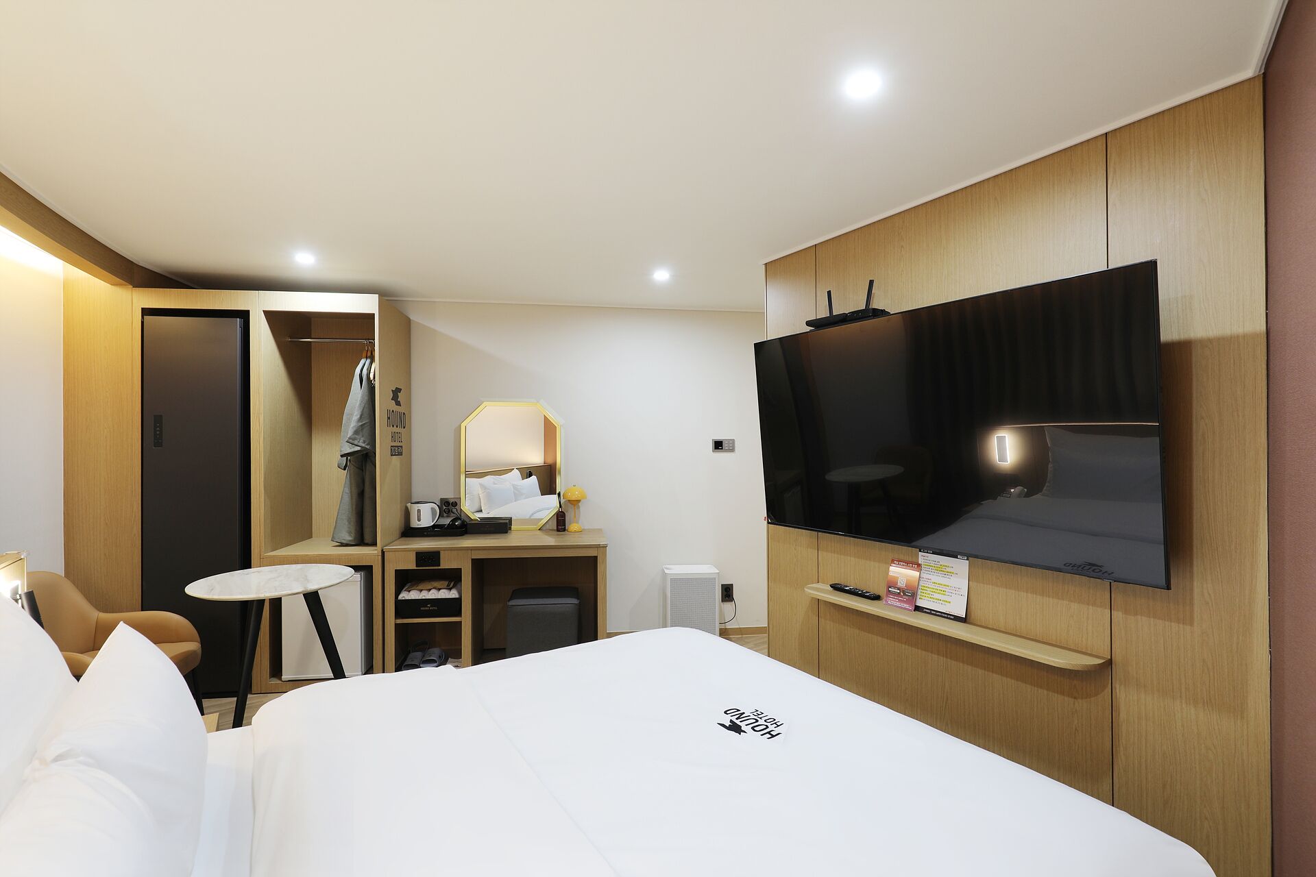 A hotel room with a bed, a TV mounted on the wall, a desk with a chair, a wardrobe, and a small table.